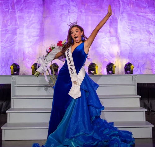 A young lady on a blue dress is holding a bouquet of flowers on her right hand while lifting her left hand up giving the appearance of excitement.